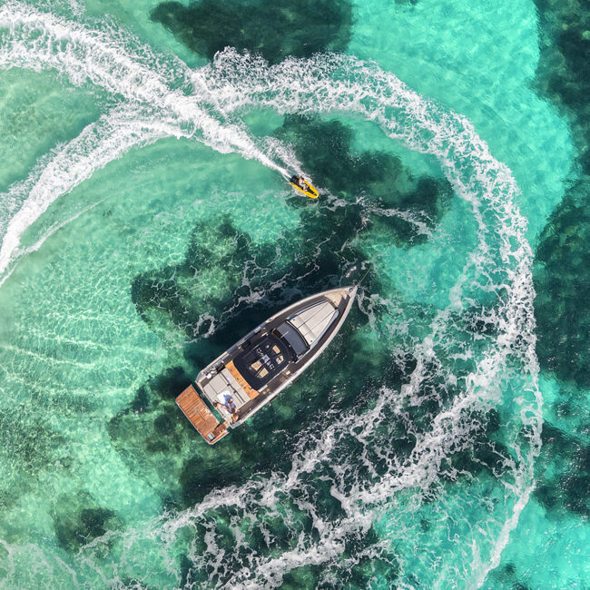 Vista aérea de yate navegando en aguas turquesas con motos de agua alrededor.