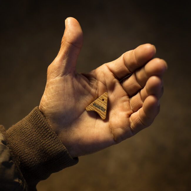 Mano sosteniendo una pequeña pieza triangular de madera sobre fondo oscuro con iluminación cálida.